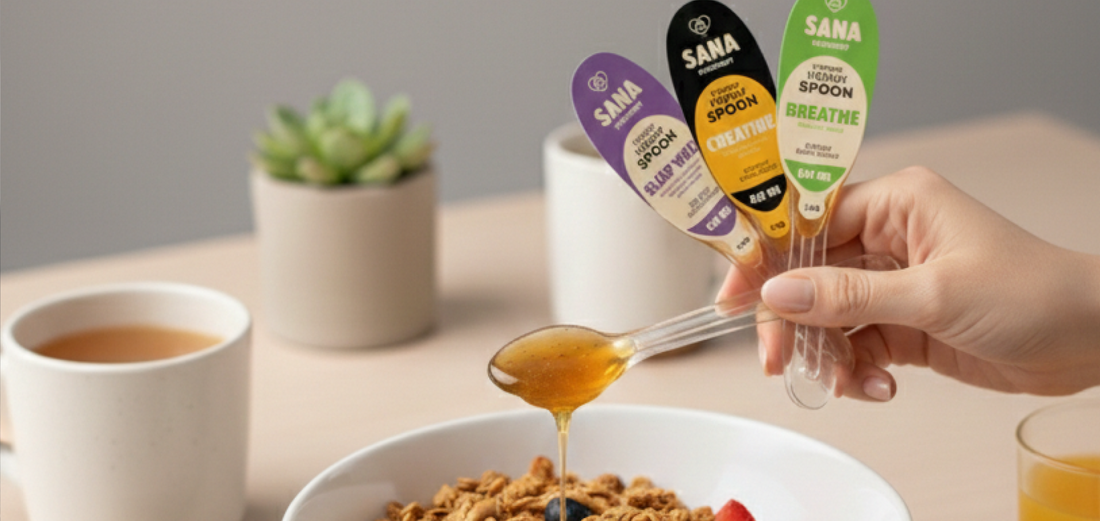 A close-up shot of a hand holding three colorful "Sana" brand honey spoons above a white bowl of yogurt, granola, strawberries, and blueberries.